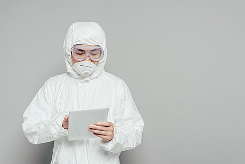 asian epidemiologist in hazmat suit and respirator mask using digital tablet on grey background