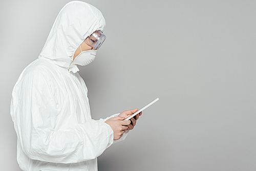 side view of asian epidemiologist in hazmat suit and respirator mask using digital tablet on grey background