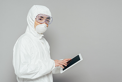 asian epidemiologist in hazmat suit and respirator mask holding digital tablet with blank screen and  on grey background