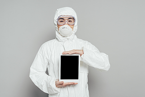 asian epidemiologist in hazmat suit and respirator mask showing digital tablet with blank screen on grey background