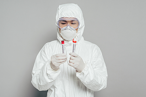 asian epidemiologist in hazmat suit and respirator mask holding test tubes with blood samples on grey background