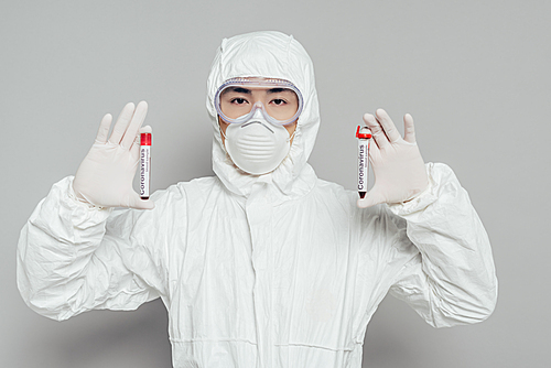 asian epidemiologist in hazmat suit and respirator mask showing test tubes with blood samples on grey background