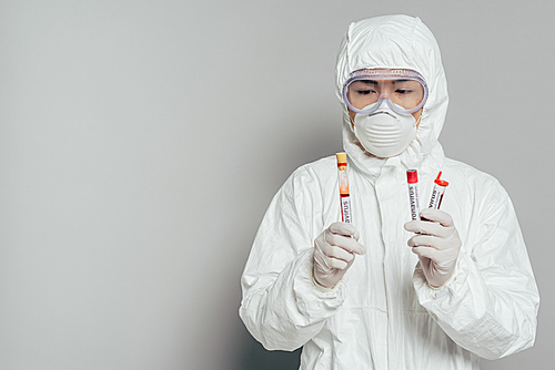 asian epidemiologist in hazmat suit and respirator mask holding test tubes with blood samples on grey background