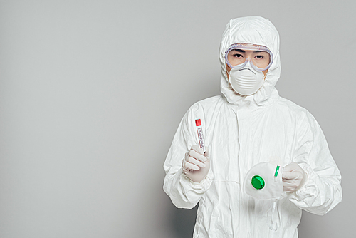 asian epidemiologist in hazmat suit  while holding respirator mask and test tube with blood sample on grey background
