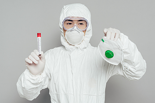 asian epidemiologist in hazmat suit holding respirator mask and test tube with blood sample on grey background