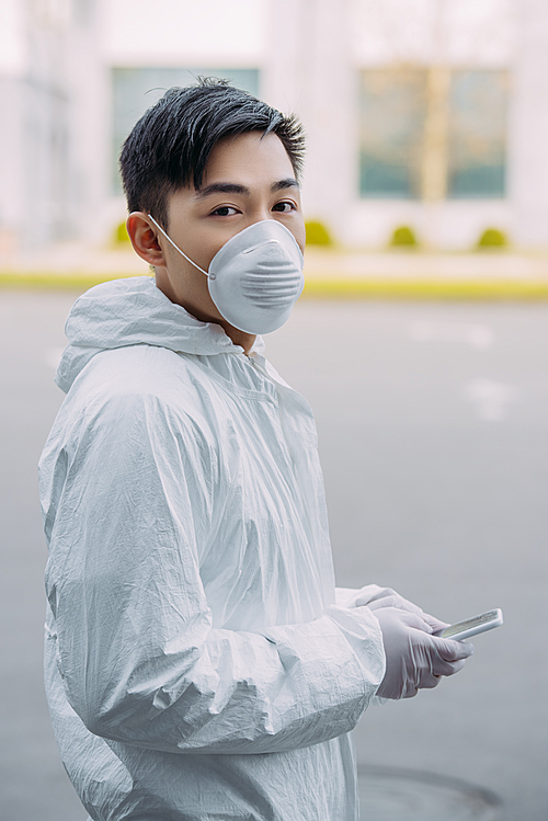 asian epidemiologist in hazmat suit and respirator mask holding smartphone and  while standing on street
