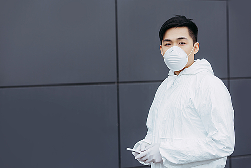 asian epidemiologist in hazmat suit and respirator mask holding smartphone and  while standing near wall