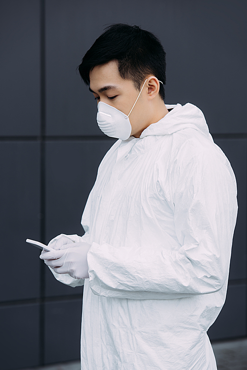 asian epidemiologist in hazmat suit and respirator mask chatting on smartphone while standing outside