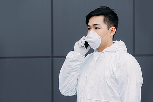 asian epidemiologist in hazmat suit and respirator mask talking on smartphone and looking away outside