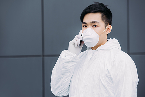 asian epidemiologist in hazmat suit and respirator mask talking on smartphone and  while standing on street