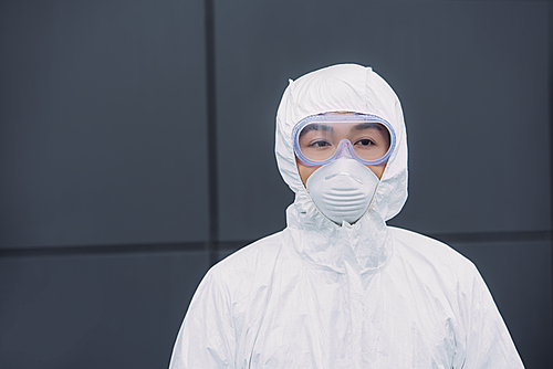 asian epidemiologist in hazmat suit and respirator mask looking away while standing near wall of building