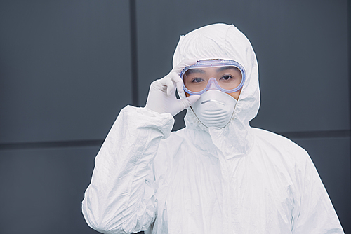 asian epidemiologist in hazmat suit and respirator mask  while standing outside and touching goggles