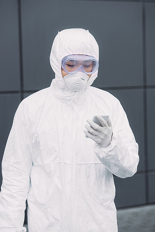 asian epidemiologist in hazmat suit and respirator mask standing outside and looking at smartphone