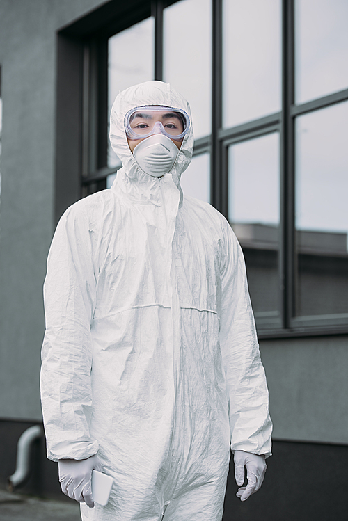 asian epidemiologist in hazmat suit and respirator mask looking away while standing near building