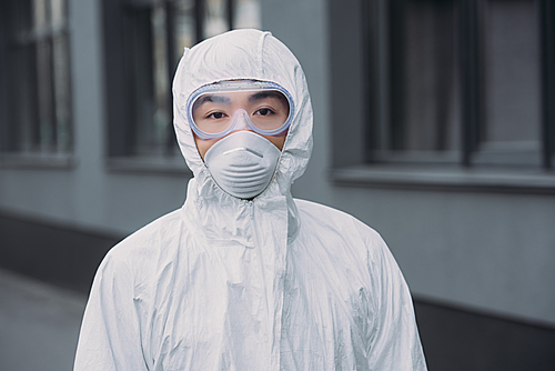 asian epidemiologist in hazmat suit and respirator mask  while standing near building