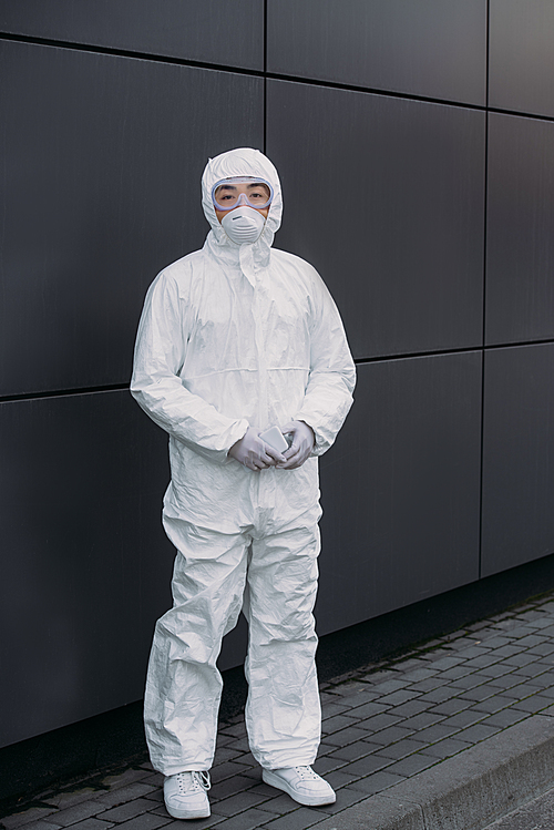 asian epidemiologist in hazmat suit and respirator mask  while standing near wall