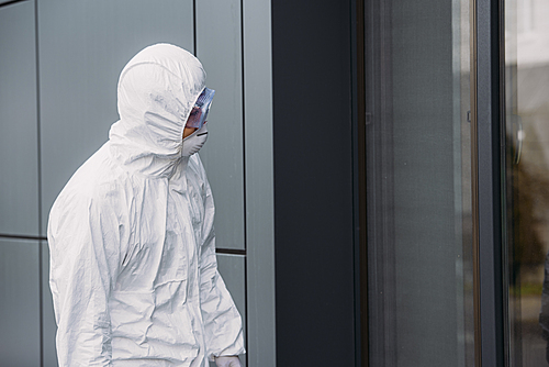 epidemiologist in hazmat suit standing outside and looking in window of building