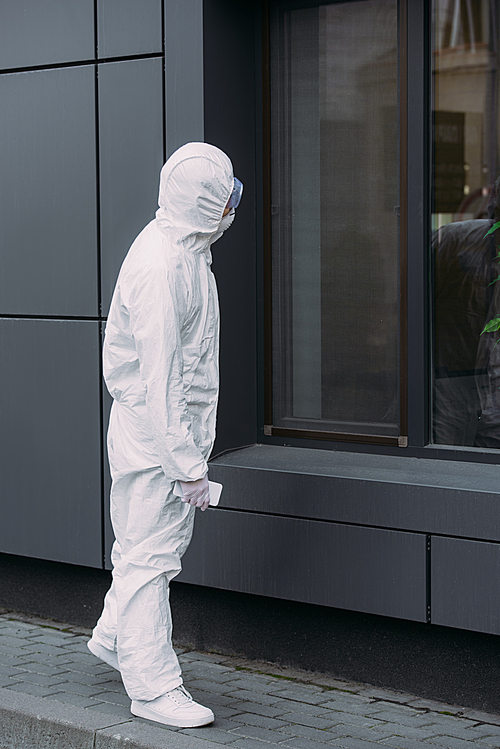 epidemiologist in hazmat suit standing on street and looking in window of building
