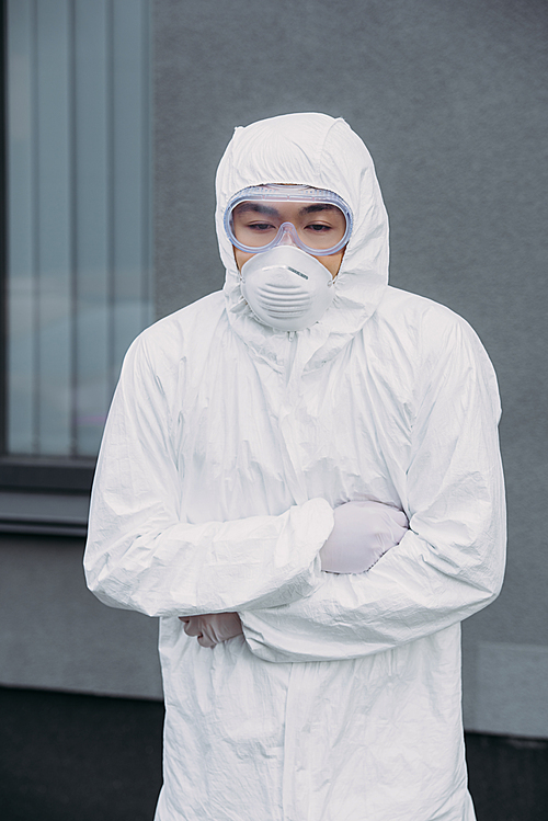 asian epidemiologist in hazmat suit and respirator mask standing with crossed arms while suffering from symptomatic abdominal pain on street