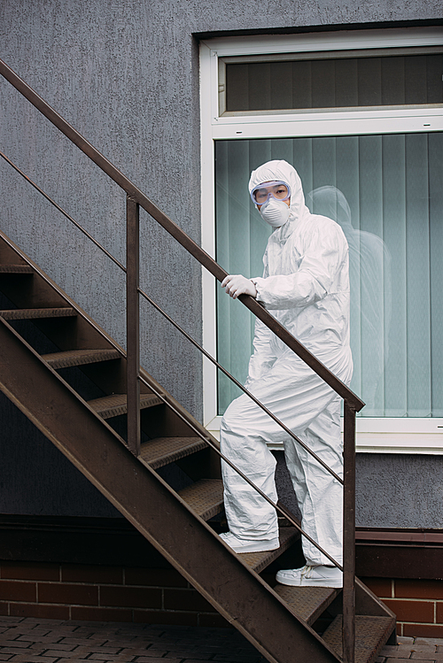 asian epidemiologist in hazmat suit and respirator mask walking upstairs outside building