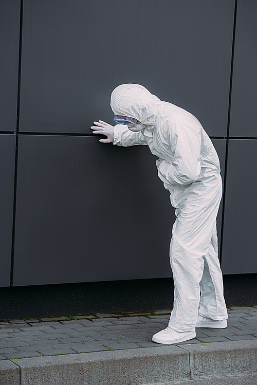 asian epidemiologist in hazmat suit leaning on wall while suffering from symptomatic abdominal pain