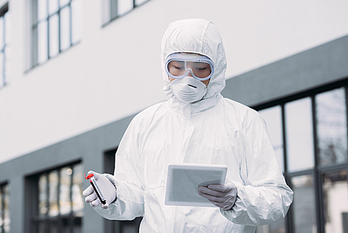 asian epidemiologist in hazmat suit and respirator mask using digital tablet while holding test tube with blood sample on street