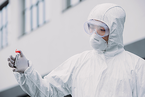 asian epidemiologist in hazmat suit and respirator mask holding test tube with blood sample while standing outdoors