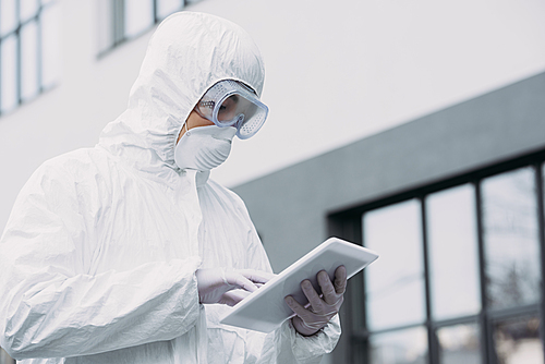 asian epidemiologist in hazmat suit and respirator mask using digital tablet while standing on street