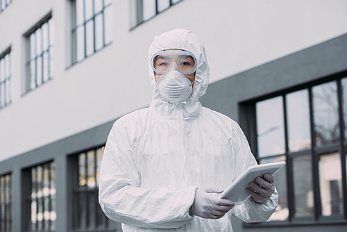 asian epidemiologist in hazmat suit and respirator mask holding digital tablet and  while standing on street