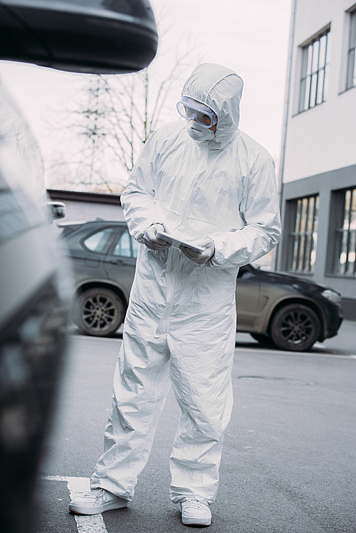 selective focus of epidemiologist in hazmat suit holding digital tablet while checking cars on parking lot