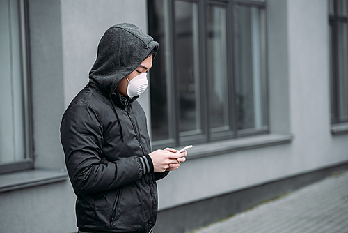 young asian man in respirator mask chatting on smartphone while standing on street