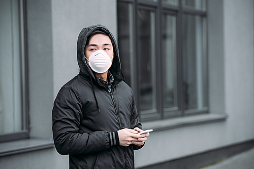 young asian man in respirator mask holding smartphone and looking away while standing on street