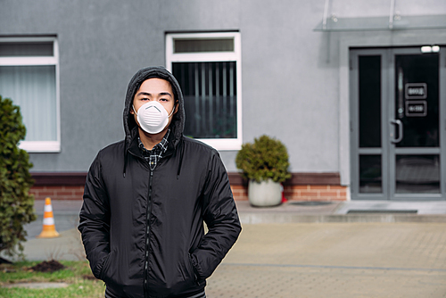 young asian man in respirator mask  and holding hands in pockets while standing on street
