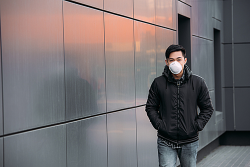 young asian man in respirator mask  while walking along wall with hands in pockets