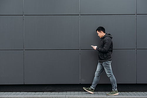side view of young asian man chatting on smartphone while walking along wall