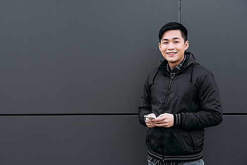 young asian man smiling at camera while standing near wall and using smartphone