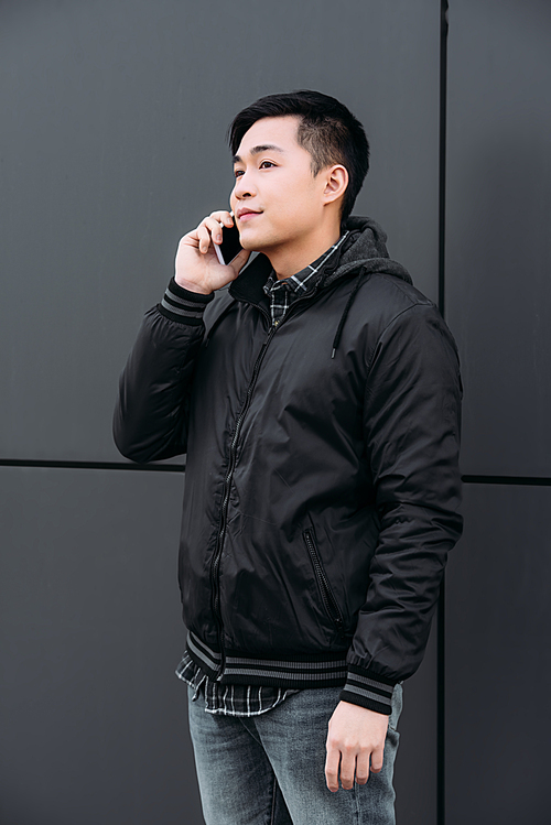 young asian man looking away while standing by wall and talking on smartphone