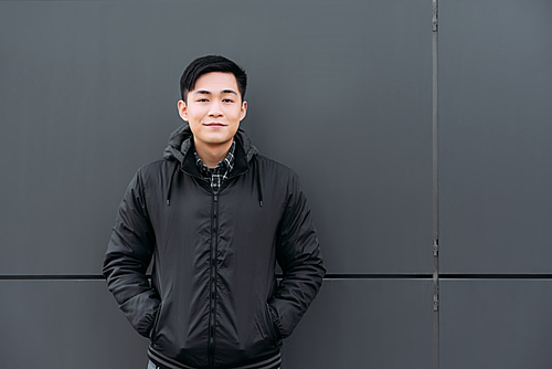young asian man smiling at camera while standing by wall and holding hands in pockets