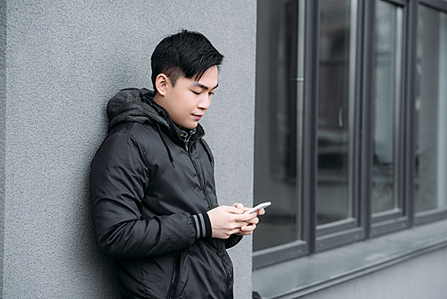 young asian man chatting on smartphone while standing by grey wall