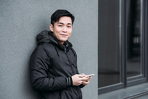 young asian man smiling at camera while standing by grey wall and chatting on smartphone