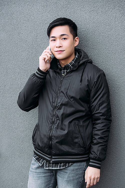 young asian man smiling while standing by grey wall, talking on smartphone and 