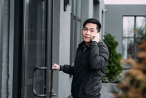 young asian man looking away while entering building and talking on smartphone
