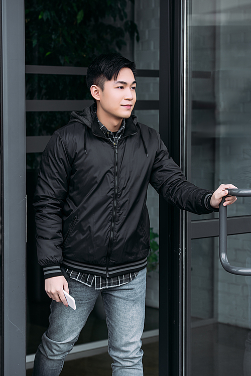young asian man holding smartphone and looking away while leaving building
