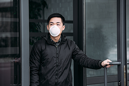 young asian man in respirator mask looking away while leaving building