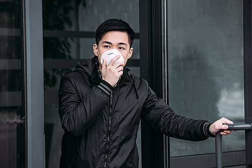 young asian man touching respirator mask while going out of building and looking away