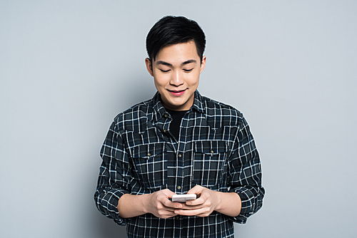 young asian man smiling while using smartphone on grey background