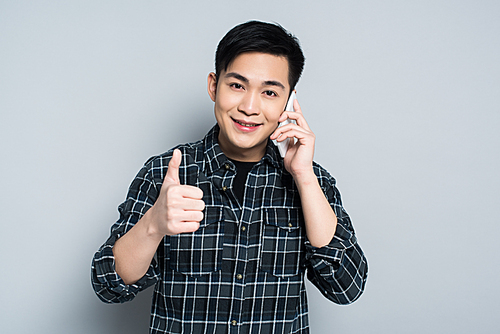 young asian man smiling at camera while talking on smartphone and showing thumb up on grey background