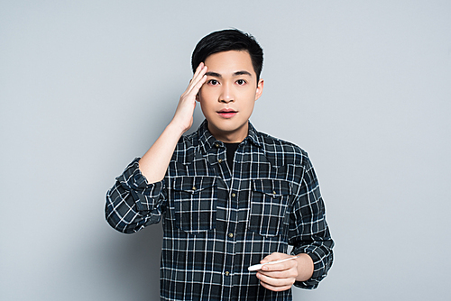 shocked asian man touching forehead and  while holding thermometer on grey background
