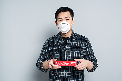 young asian man in respirator mask showing warning card with coronavirus inscription on grey background