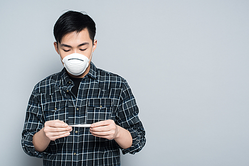 young asian man in respirator mask looking at thermometer on grey background